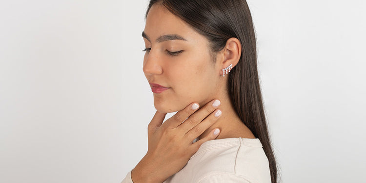 Frau mit glatten brünetten Haaren trägt funkelnde Ohrschieber und ein weißes Oberteil, Hand elegant am Hals positioniert, vor hellem Hintergrund