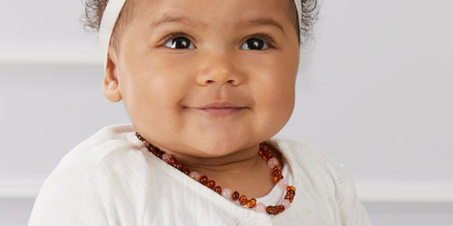 Lächelndes Baby mit lockigen Haaren trägt braune Bernsteinkette und kleine Ohrstecker