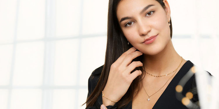Lächelnde Frau mit langen braunen Haarenin schwarzem Top trägt goldene Layering-Halsketten mit Stern-Anhänger und goldenes Armband 