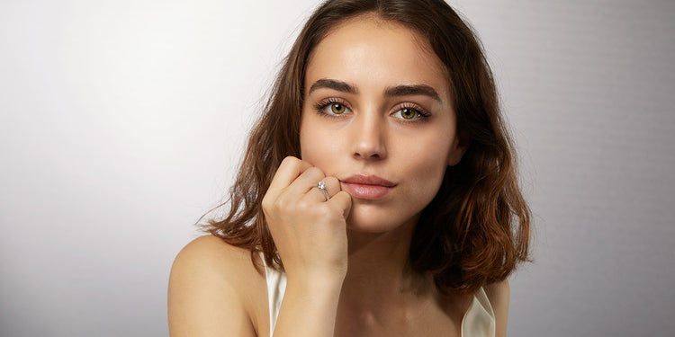 Junge Frau mit braunen Haaren trägt einen eleganten Solitär-Ring, Hand am Kinn posierend vor grauem Hintergrund