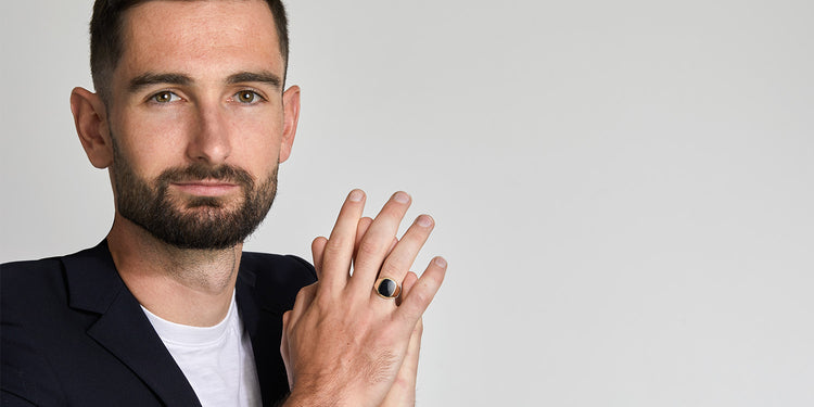 Eleganter Mann mit Bart in schwarzem Blazer präsentiert silbernen Siegelring mit schwarzem Stein an der Hand