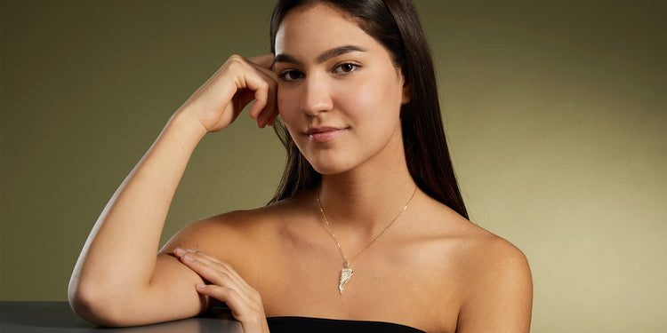 Junge Frau mit langen dunklen Haaren trägt eine goldene Halskette mit Flügel-Anhänger und ein schwarzes schulterfreies Top